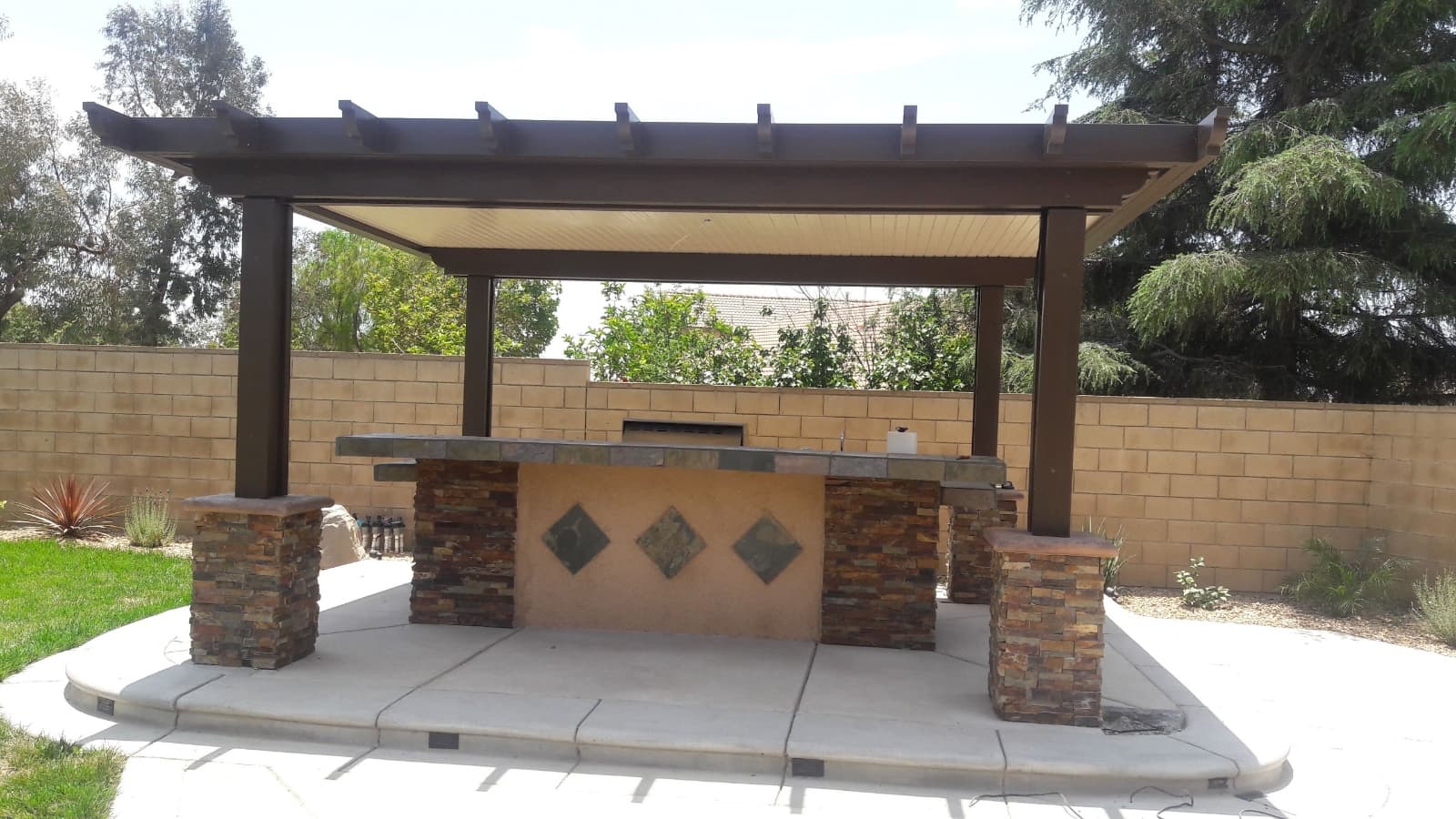 Outdoor stone bar with diamond accents under a dark brown pergola in a backyard.