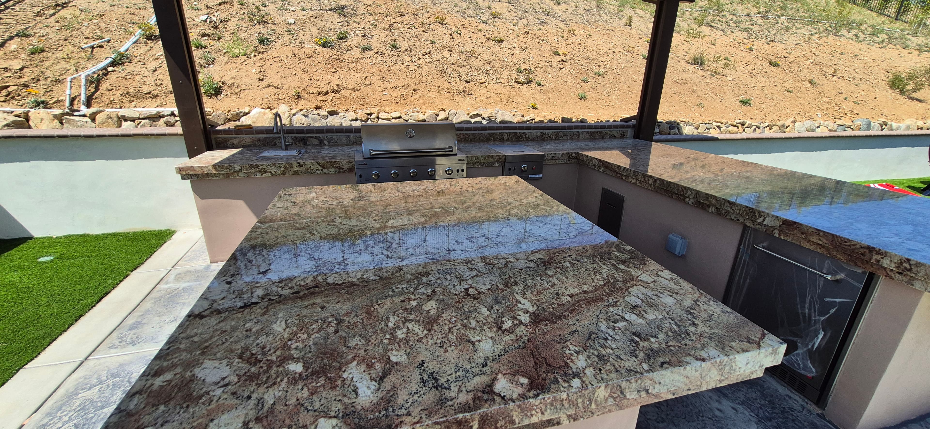 Polished granite outdoor kitchen with a stainless steel grill and sink under a dark pergola.