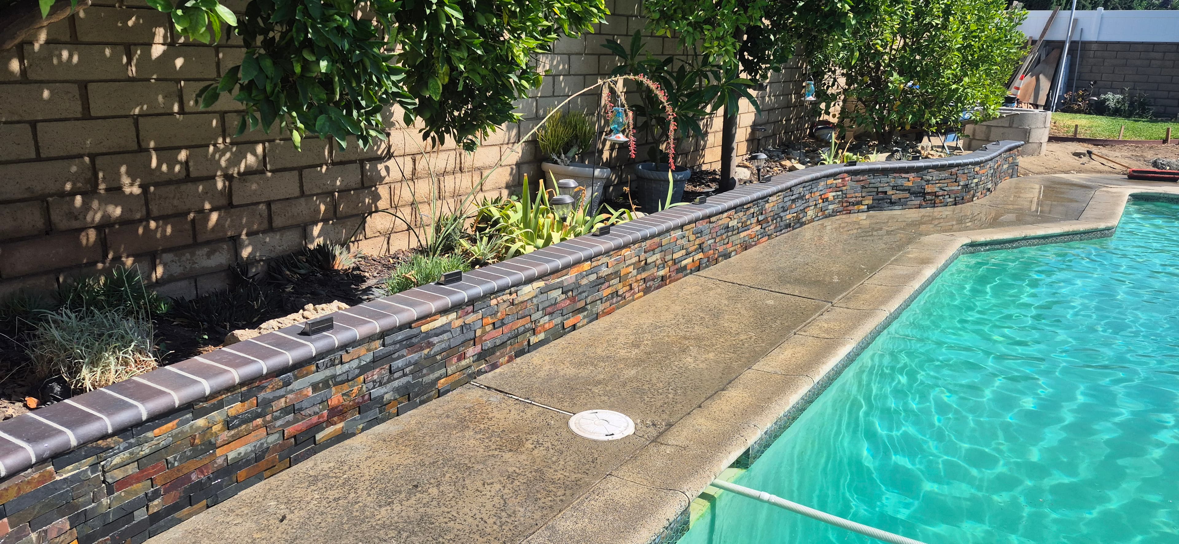 Stacked stone retaining wall with dark coping separates a lush garden from a turquoise pool.
