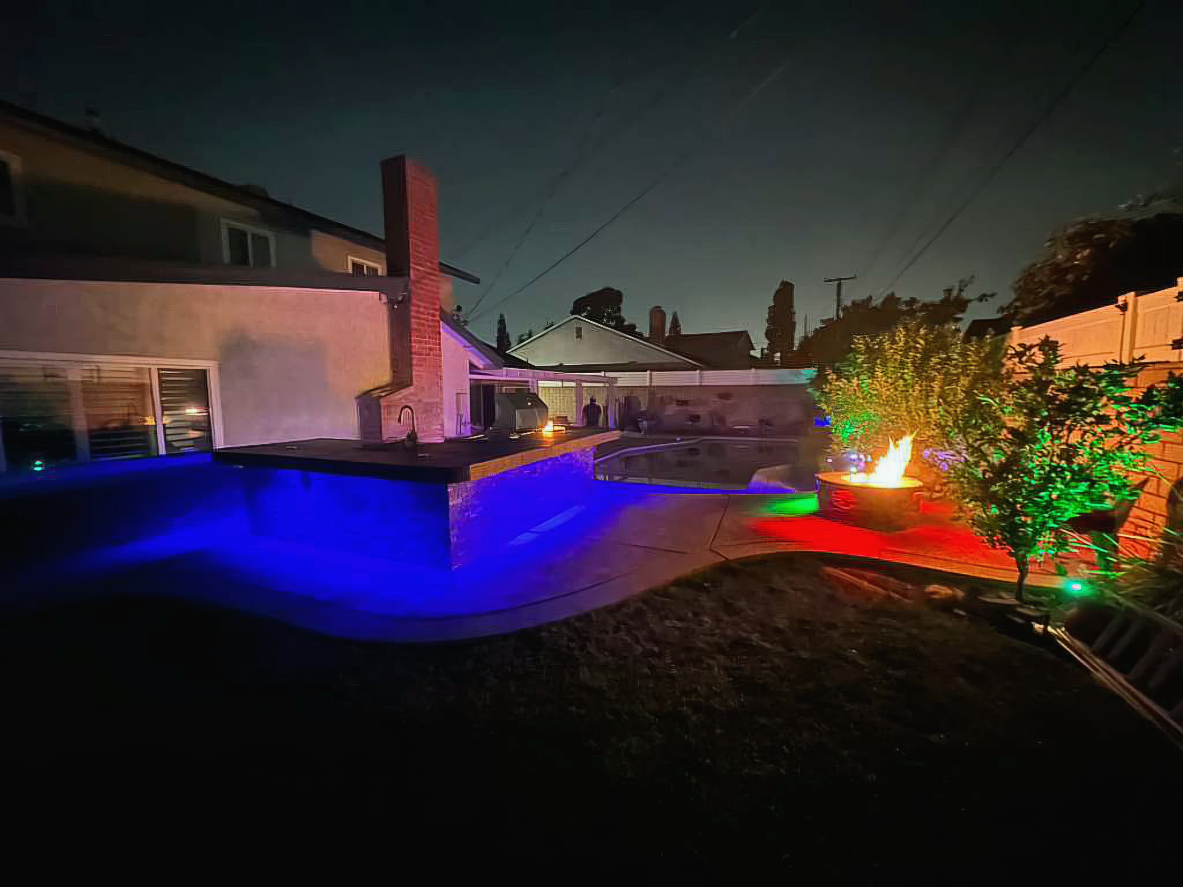 Backyard at night with blue-lit outdoor kitchen, glowing fire pit, and green-illuminated trees.
