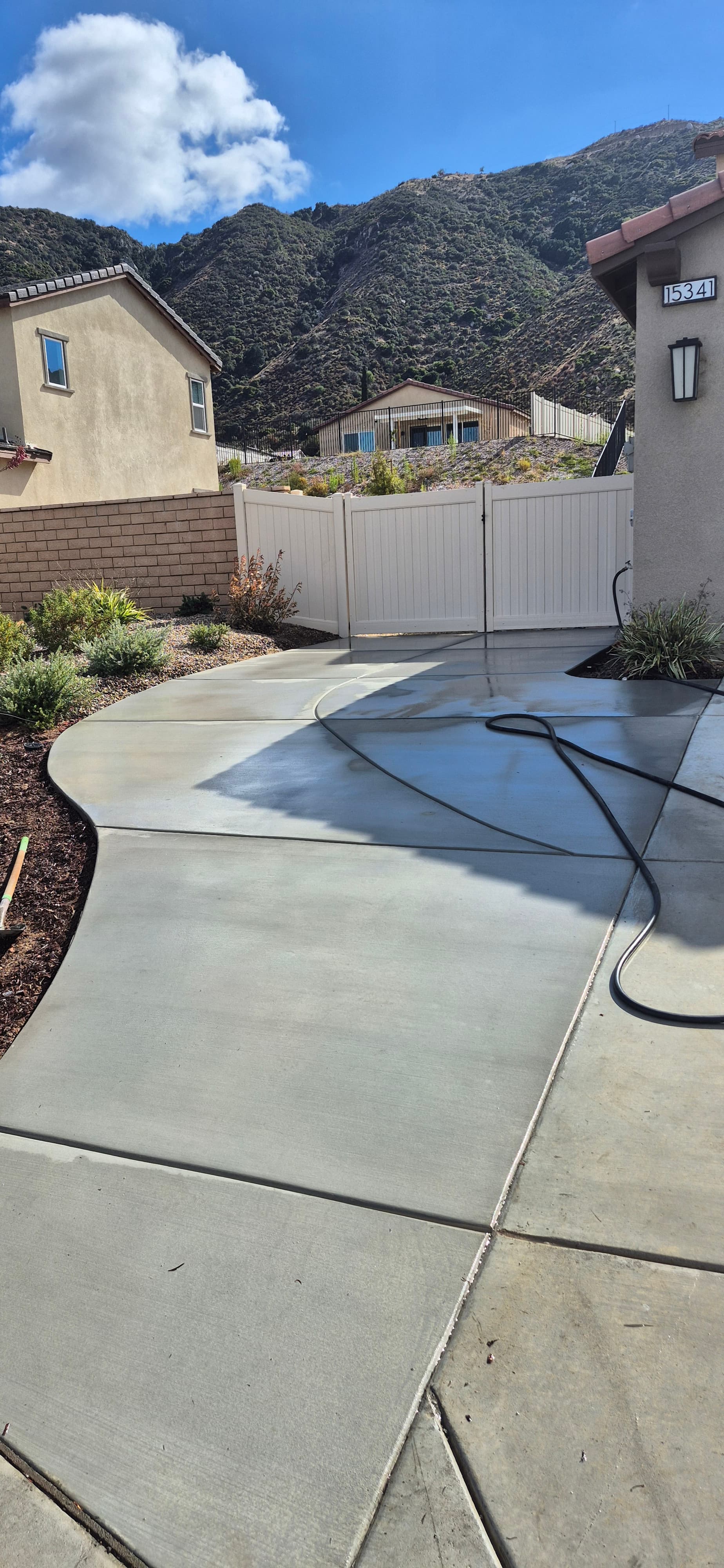 Wet concrete driveway with a black hose leading to a white gate and large mountains.