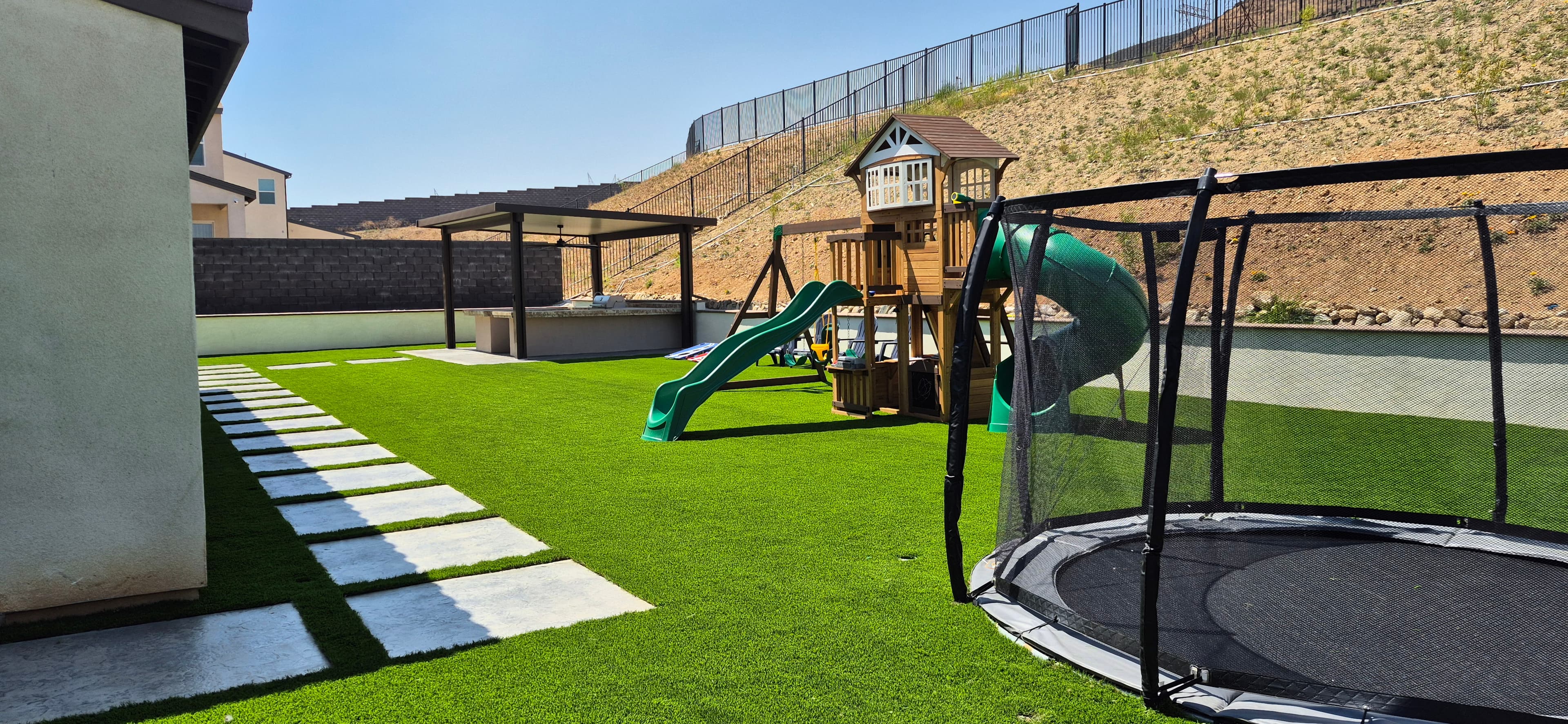 Backyard featuring artificial turf, a stone path, a wooden playset, and a large trampoline.