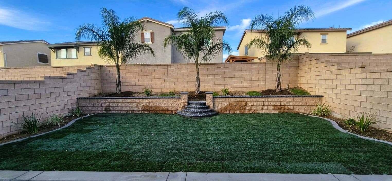Landscaped backyard featuring a green lawn, tiered stone retaining wall, and three palm trees.
