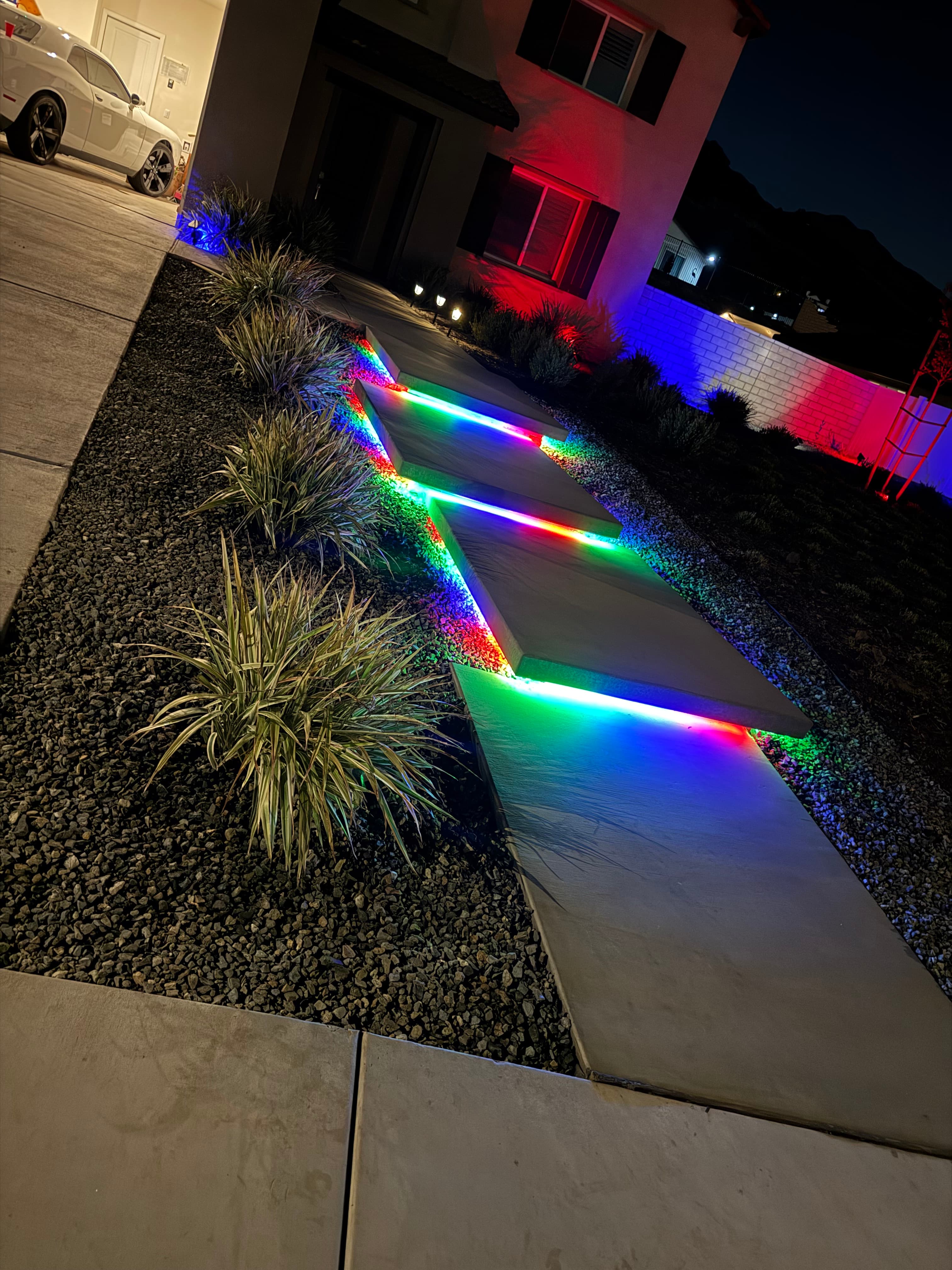 Modern concrete walkway steps glowing with vibrant rainbow LED lights at night in gravel landscaping.