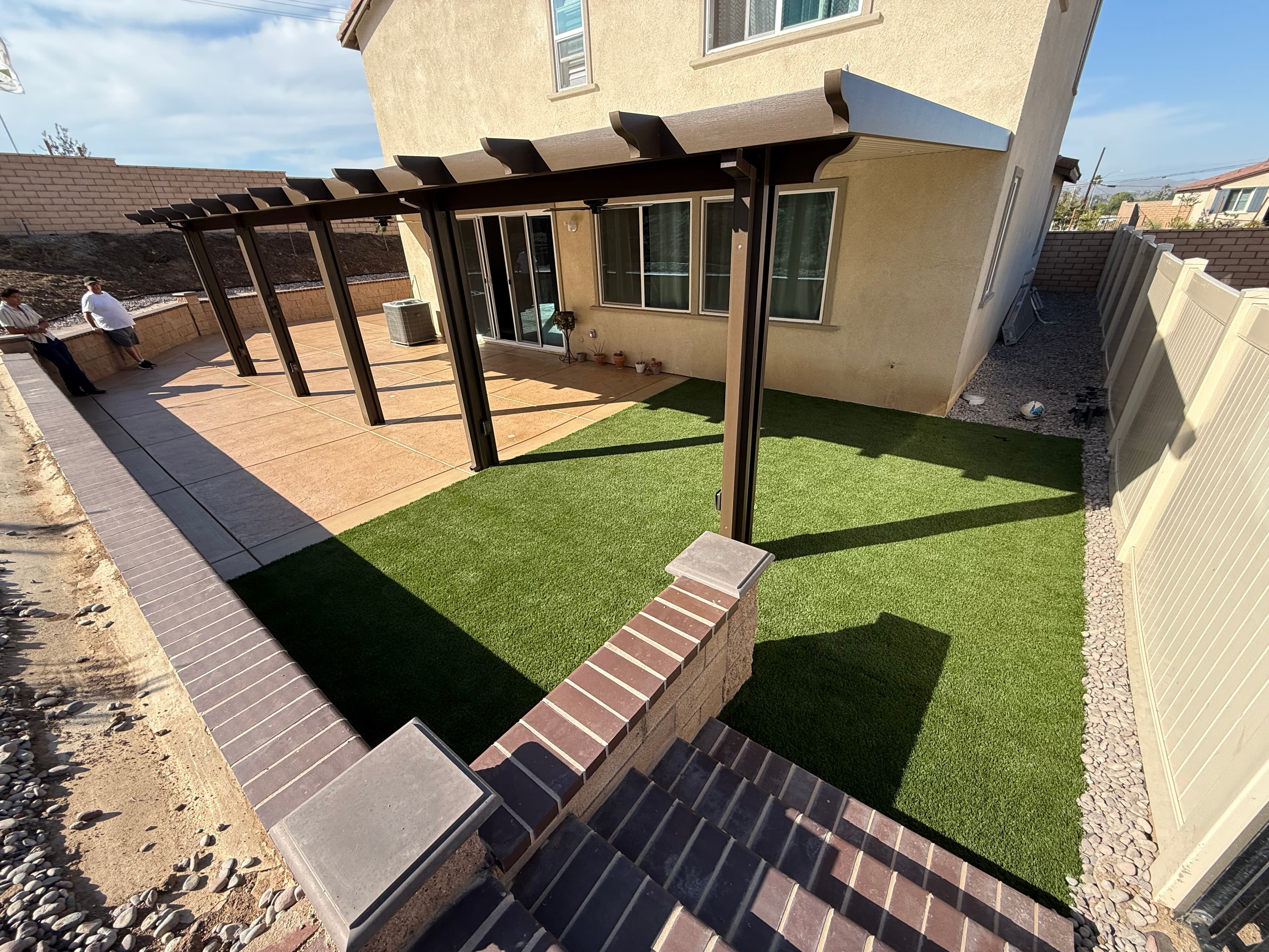 Brown metal pergola shading a backyard with artificial turf, concrete patio, and brick wall.