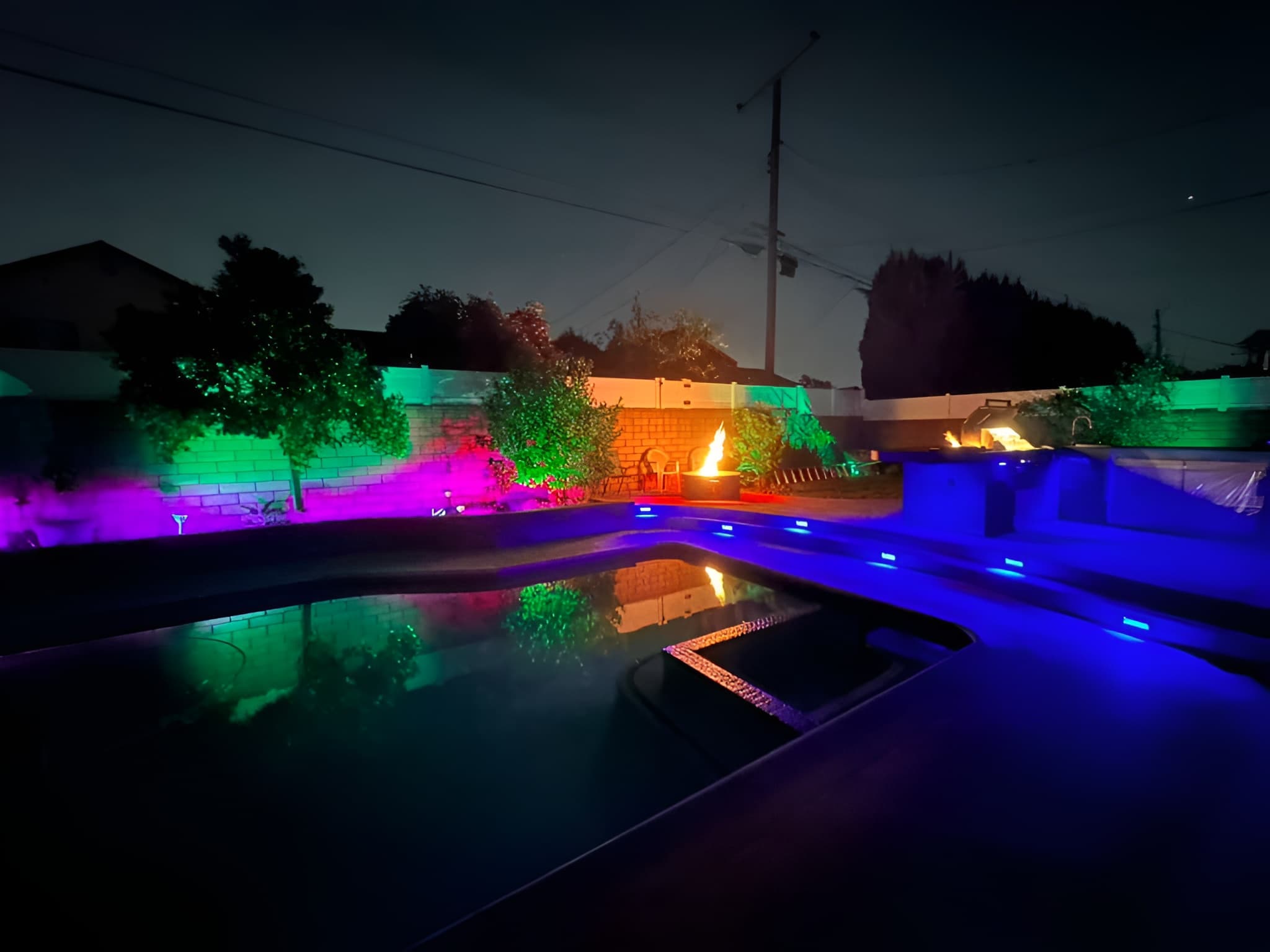 Nighttime backyard pool area featuring colorful LED lighting, a fire pit, and outdoor kitchen.