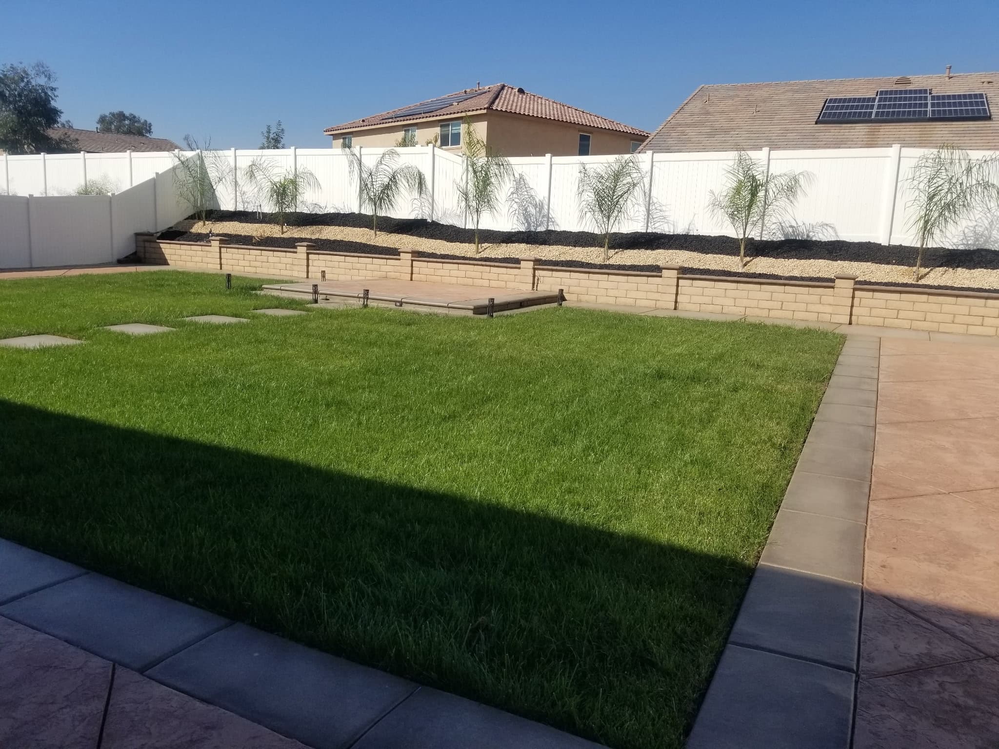 Green lawn and tiered retaining wall with palm trees against a white backyard fence.
