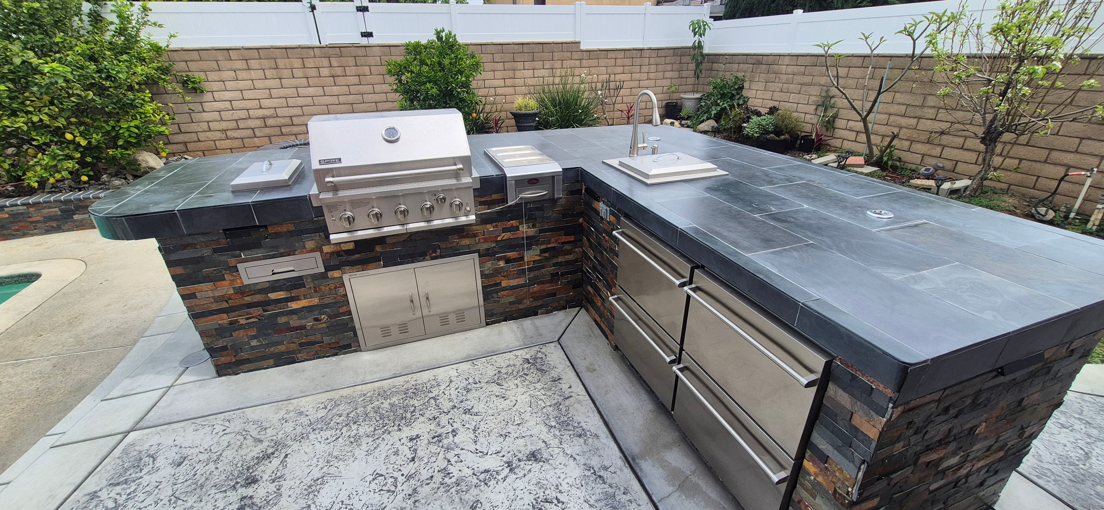 Modern outdoor kitchen island with a built-in stainless steel grill, sink, and dark tile countertops.