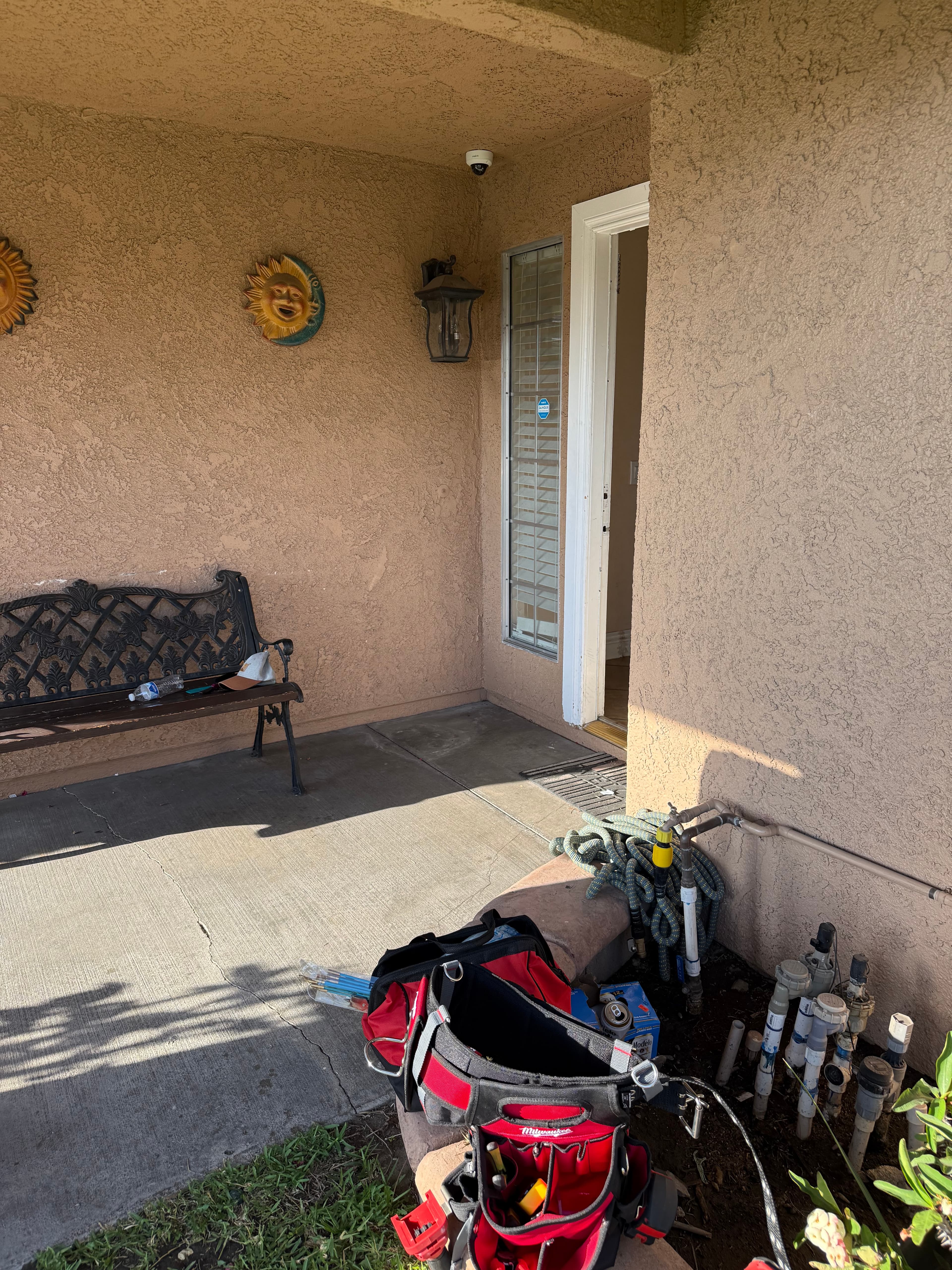Tan stucco porch with black bench, open door, and red tool bag near irrigation pipes.